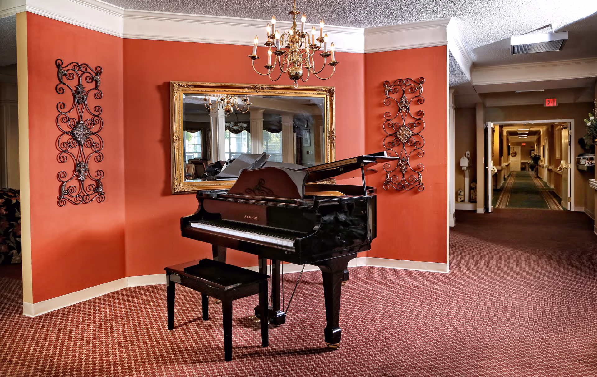 A black grand piano and bench sit in a carpeted lobby area with red walls, decorative metal wall art, a large gold-framed mirror and a chandelier.