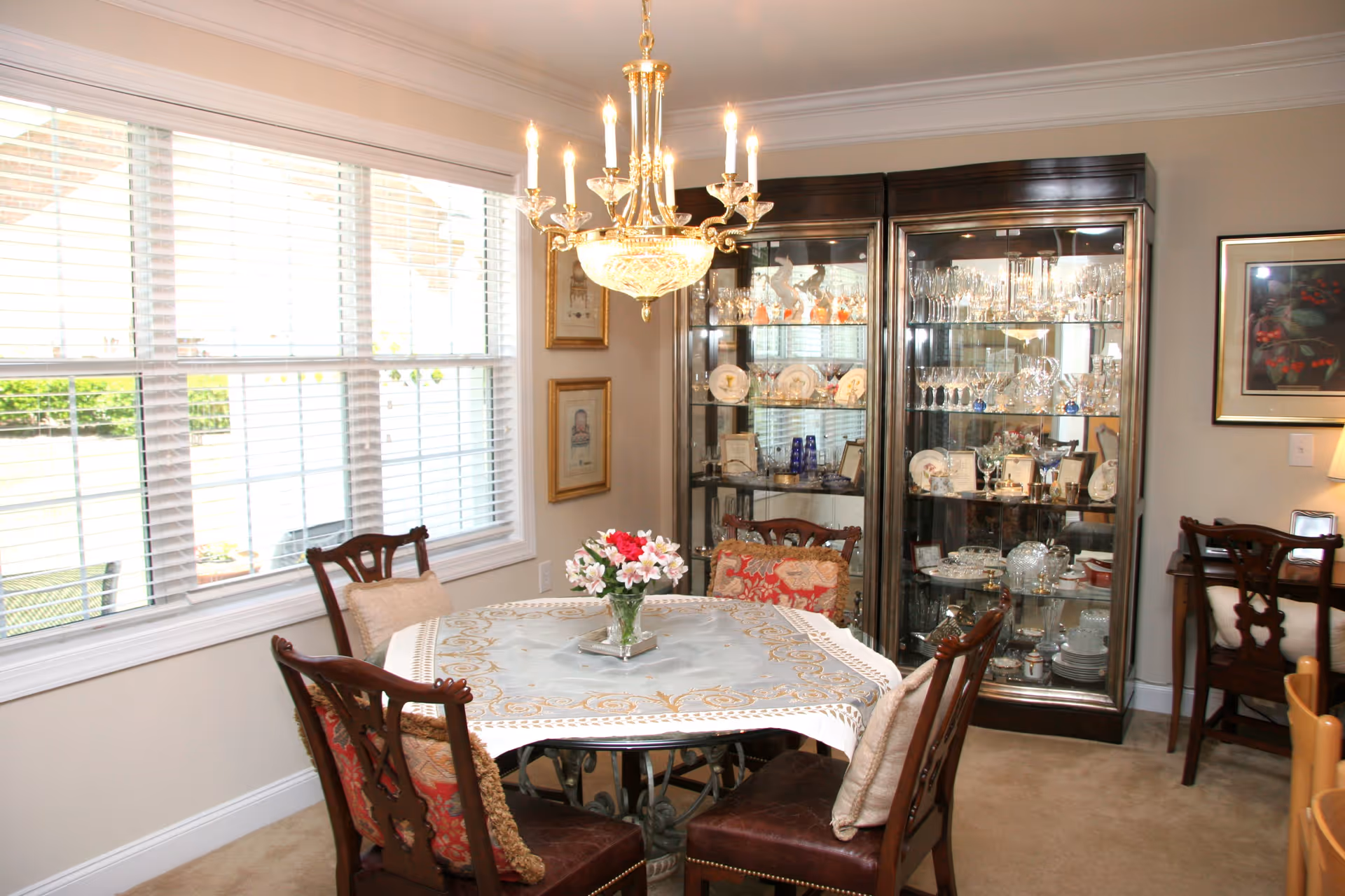 A dining room with a round table covered by a decorative tablecloth and a vase of flowers in the center. Four wooden chairs with cushions surround the table. Behind the table, there is a glass display cabinet filled with glassware and decorative plates. A chandelier with lit candles hangs above the table. Large windows with white blinds allow natural light into the room.