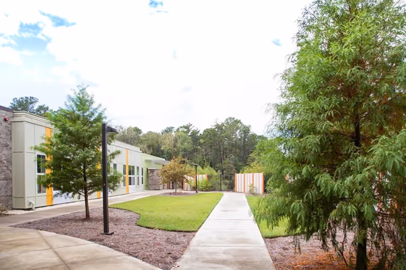 Outdoor walkway surrounded by green grass, trees, and shrubs leading to a modern building with large windows and stone accents under a partly cloudy sky.