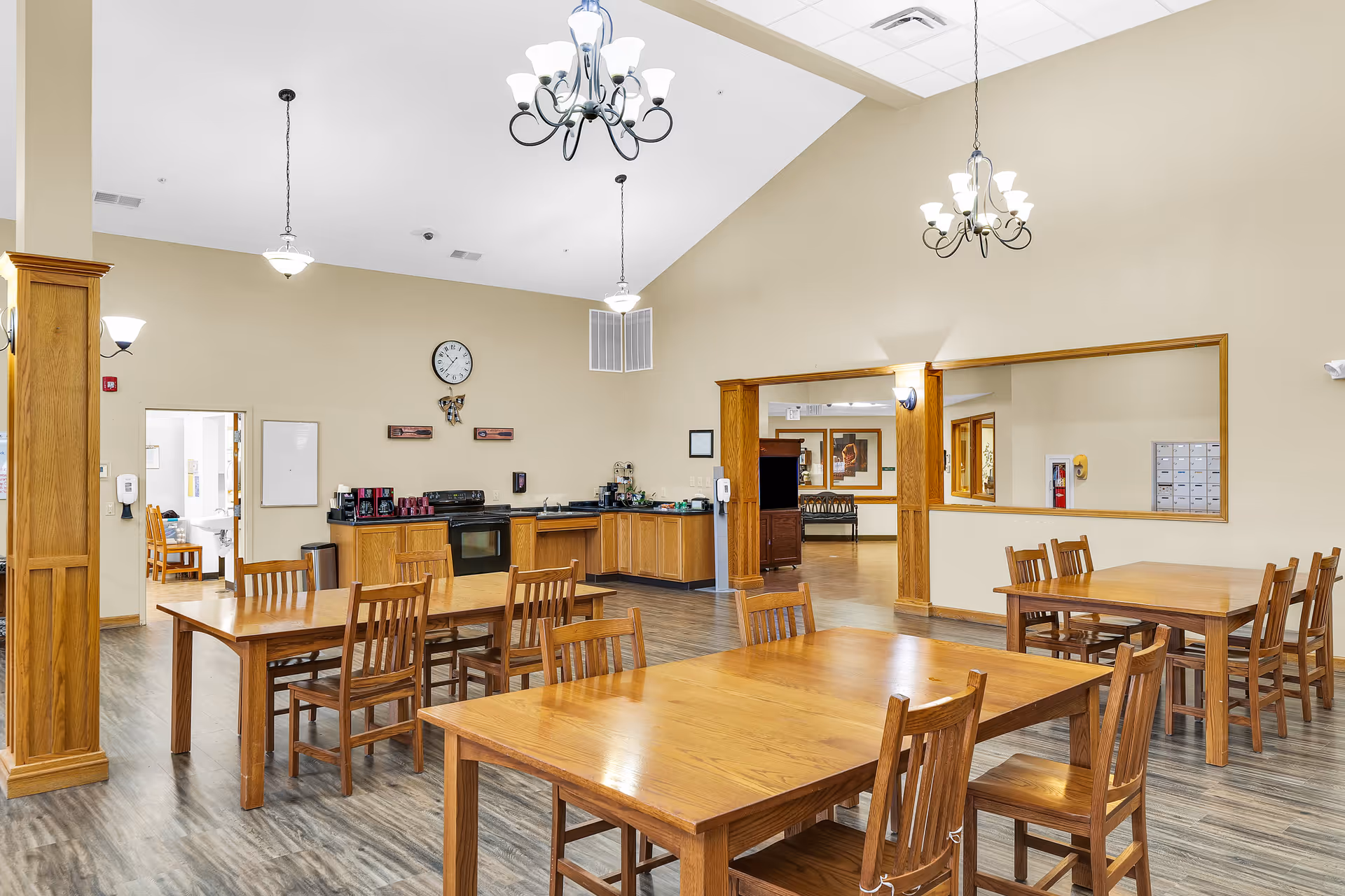 A spacious dining area in The Glenwood Assisted Living of Mahomet featuring wooden tables and chairs arranged neatly on a wood-patterned floor. The room has high ceilings with multiple chandeliers and pendant lights. In the background, there is a kitchen area with wooden cabinets, a stove, coffee makers, and a clock on the wall. The walls are painted beige, and there is a large window opening to an adjacent room.