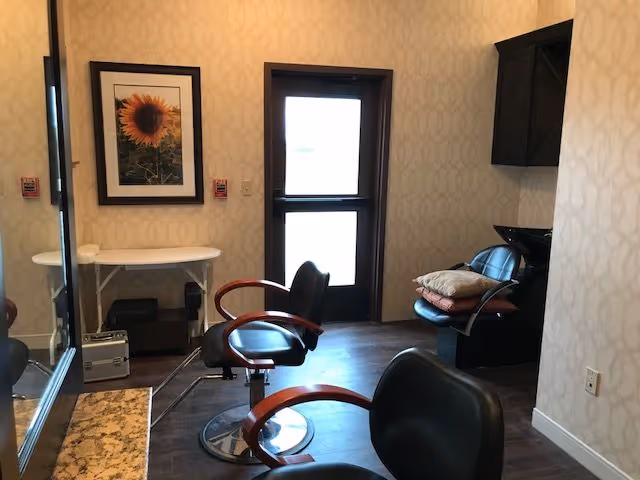 Small salon-style room with black styling chairs, a wash basin with stacked towels, a wall mirror, framed sunflower artwork, and a door.