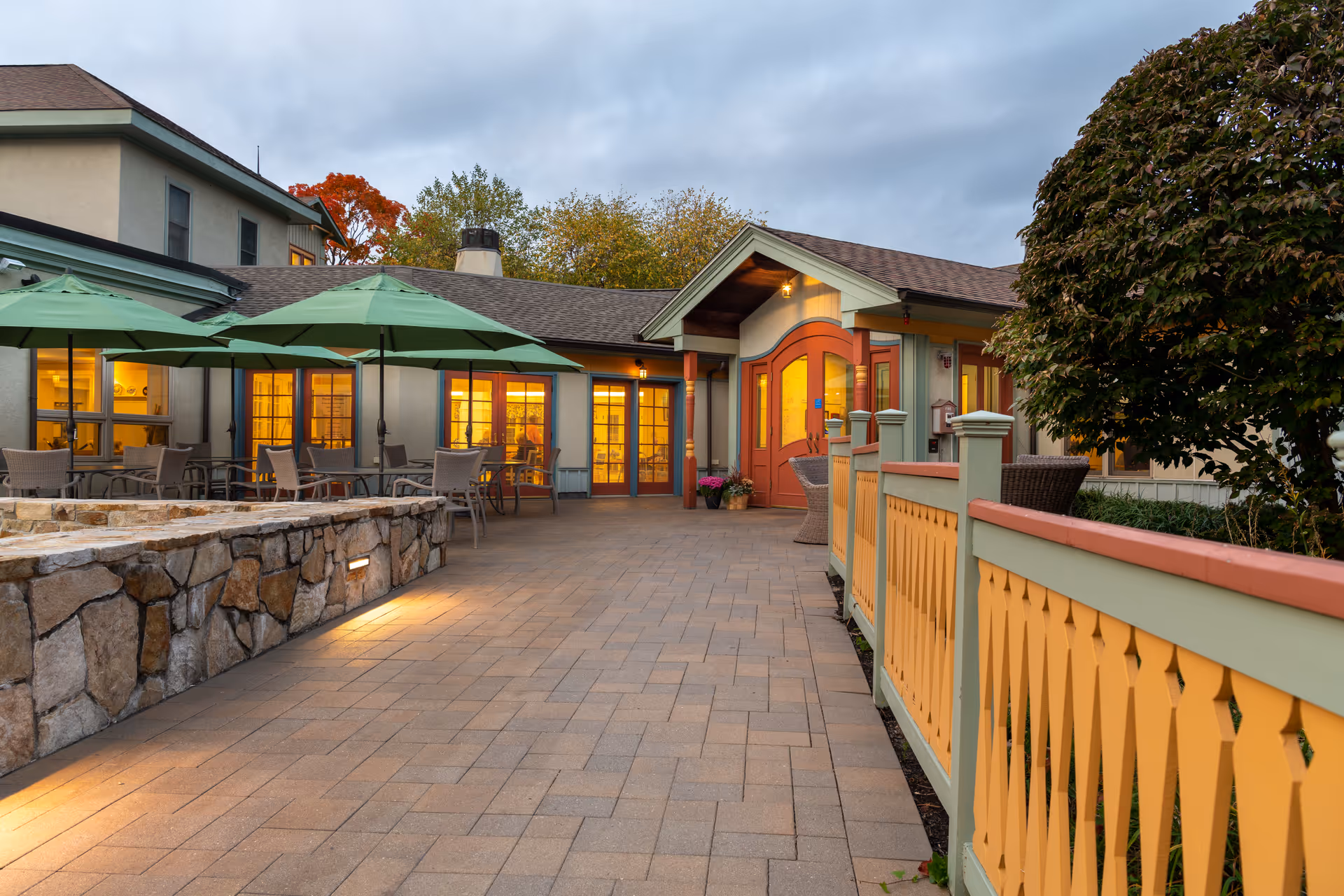 Outdoor patio area of a senior living facility with stone walls, green umbrellas over tables and chairs, a paved walkway, and a building entrance with warm lighting and a decorative fence.