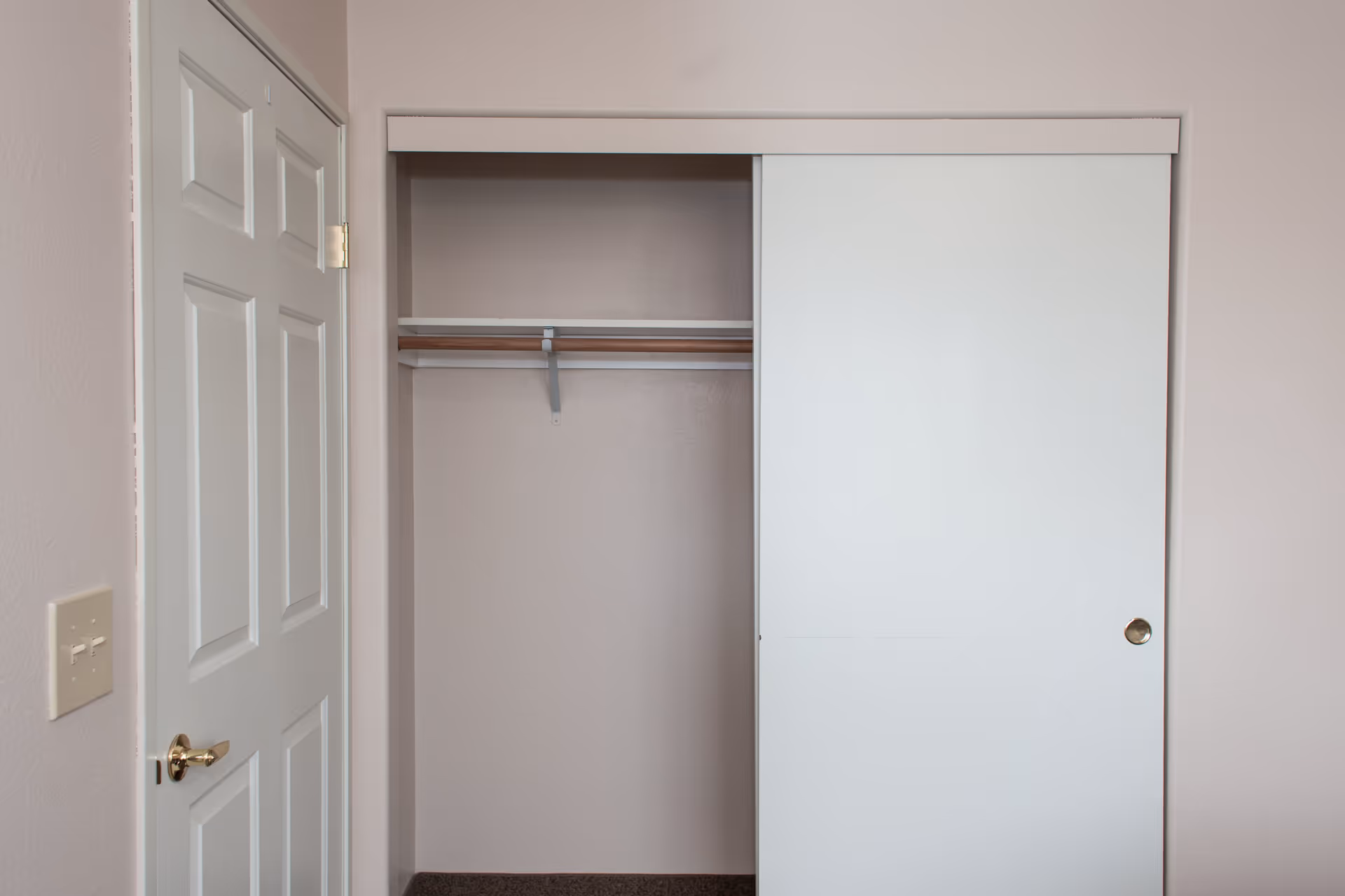 An empty closet with a sliding white door partially open, revealing a wooden hanging rod and a shelf above it. To the left of the closet is a closed white door with a gold-colored handle and a light switch on the wall beside it. The walls are painted a light beige color.
