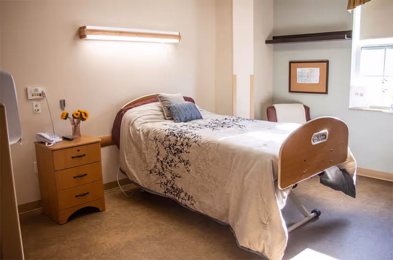 A single hospital-style bed with a beige and blue patterned blanket in a well-lit room. Next to the bed is a wooden nightstand with a telephone, remote control, and a vase with sunflowers. There is a chair with a cushion near a window with a small shelf above it. The room has beige walls and a brown floor.