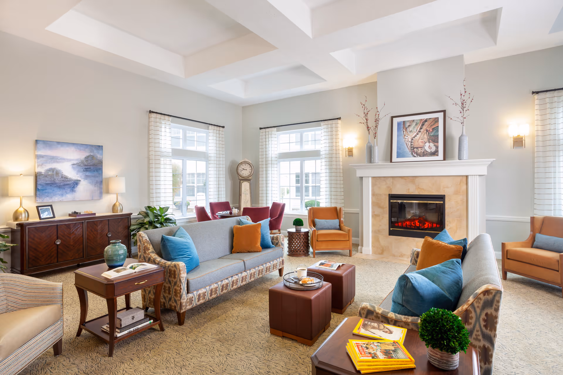 A bright and spacious living room with two patterned sofas adorned with blue and orange pillows, two brown armchairs, a fireplace with a framed artwork above it, a grandfather clock, a wooden sideboard with lamps and a painting, and several small tables with books and decorative plants. Large windows with white curtains allow natural light to fill the room.