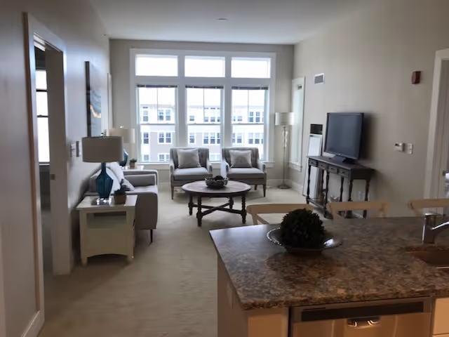 A bright living room area in an independent living community featuring a large window with multiple panes, two armchairs with cushions, a round wooden coffee table with a decorative centerpiece, a flat-screen TV on a wooden console table, and a beige sofa with a side table and lamp. In the foreground, there is a kitchen island with a decorative plant and a sink.