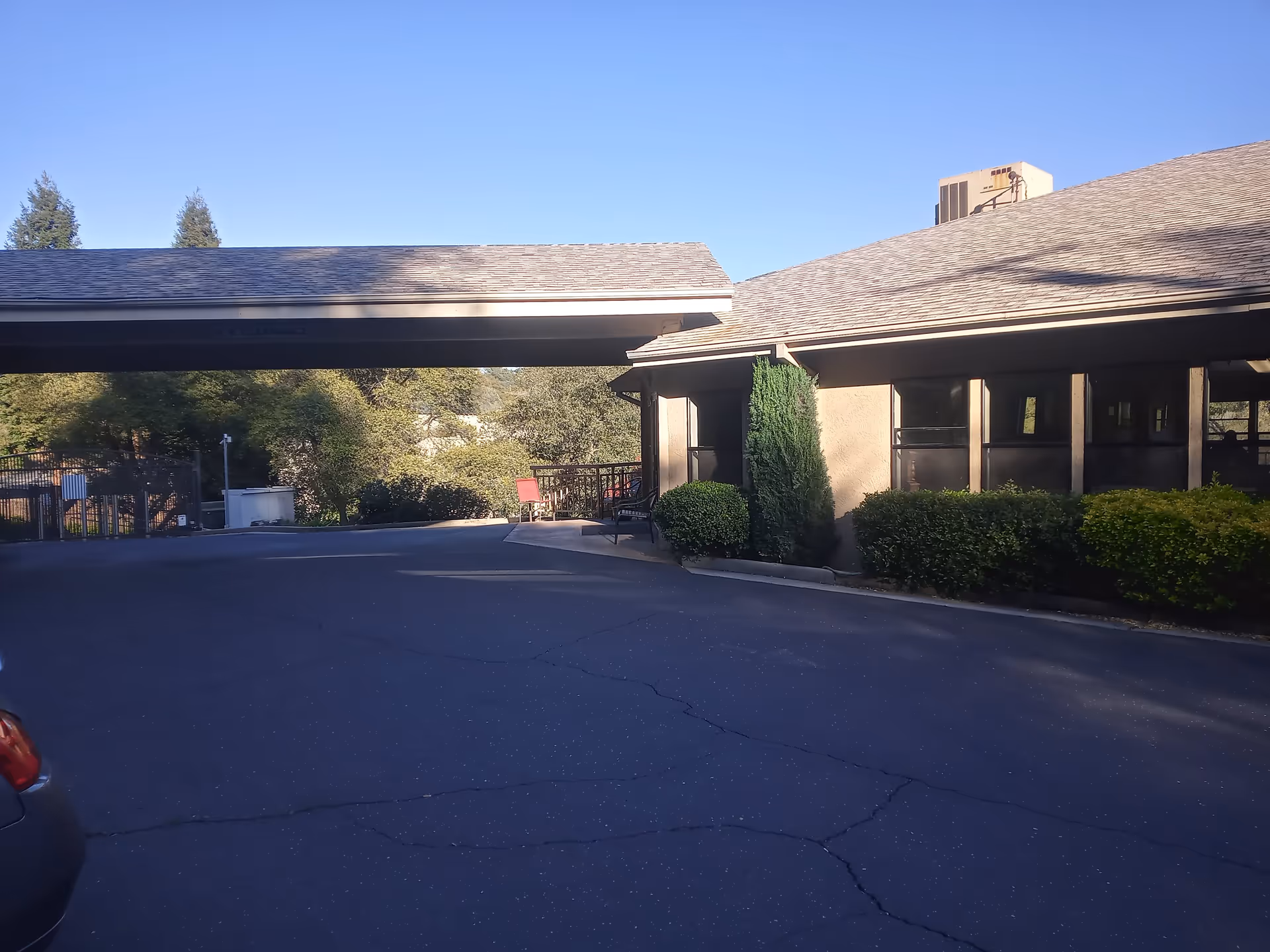 Exterior view of a senior living facility building with a covered driveway, surrounded by bushes and trees under a clear blue sky.