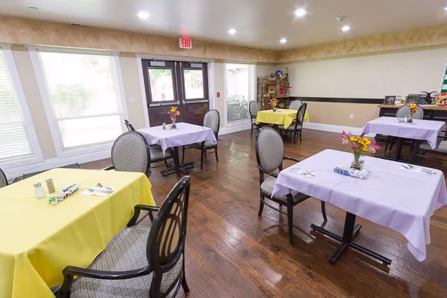 Bright dining room with multiple tables covered in pastel tablecloths and chairs arranged on a wood floor near large windows and an exit door.