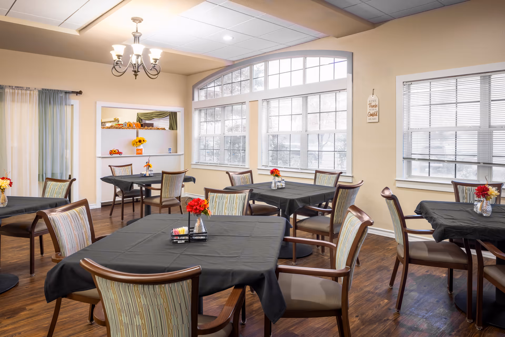 A bright dining room with several tables covered in black tablecloths, each adorned with small vases of red and yellow flowers. The room has large windows with white blinds, wooden floors, and beige walls. A chandelier hangs from the ceiling, and there is a small serving window with shelves displaying decorative items and sunflowers.