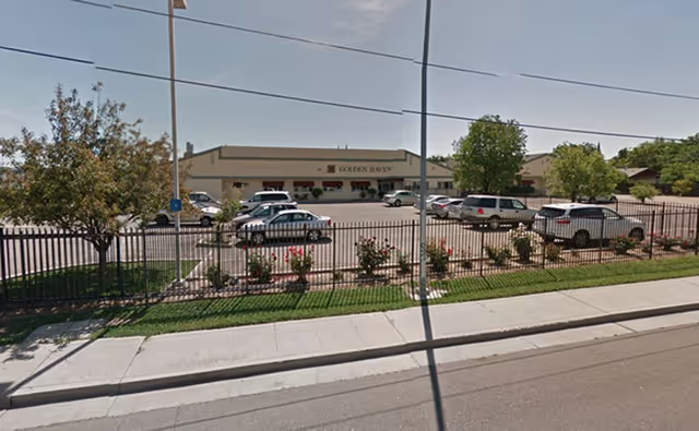 Exterior view of the Golden Haven facility showing a parking lot with several cars, a fenced garden area with small trees and shrubs, and the building in the background under a clear sky.