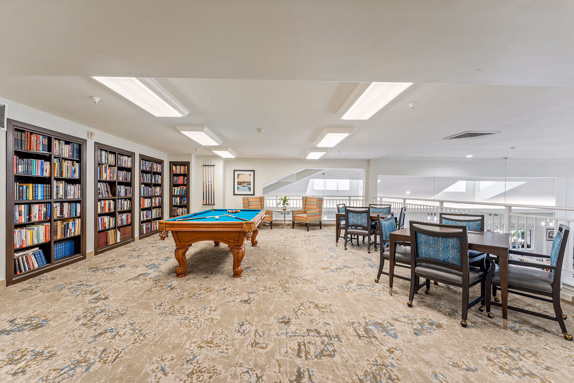 Bright communal lounge with a pool table, bookshelves, and tables and chairs overlooking an interior railing.