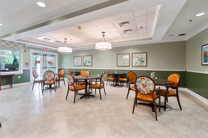 A bright dining room with several round tables and chairs featuring floral upholstery and orange seats. The walls are painted green with white trim and decorated with framed floral artwork. Two pendant lights hang from the ceiling, and there are large windows and glass doors letting in natural light.