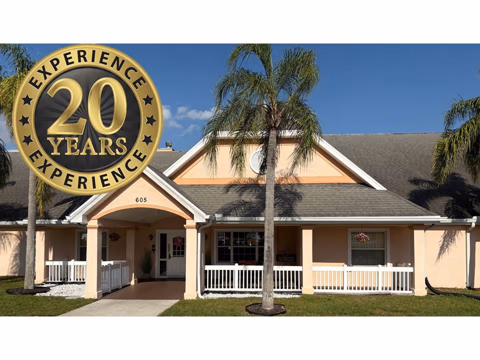 Front exterior of a single-story peach-colored senior living building with a covered porch, white railing, and palm trees.