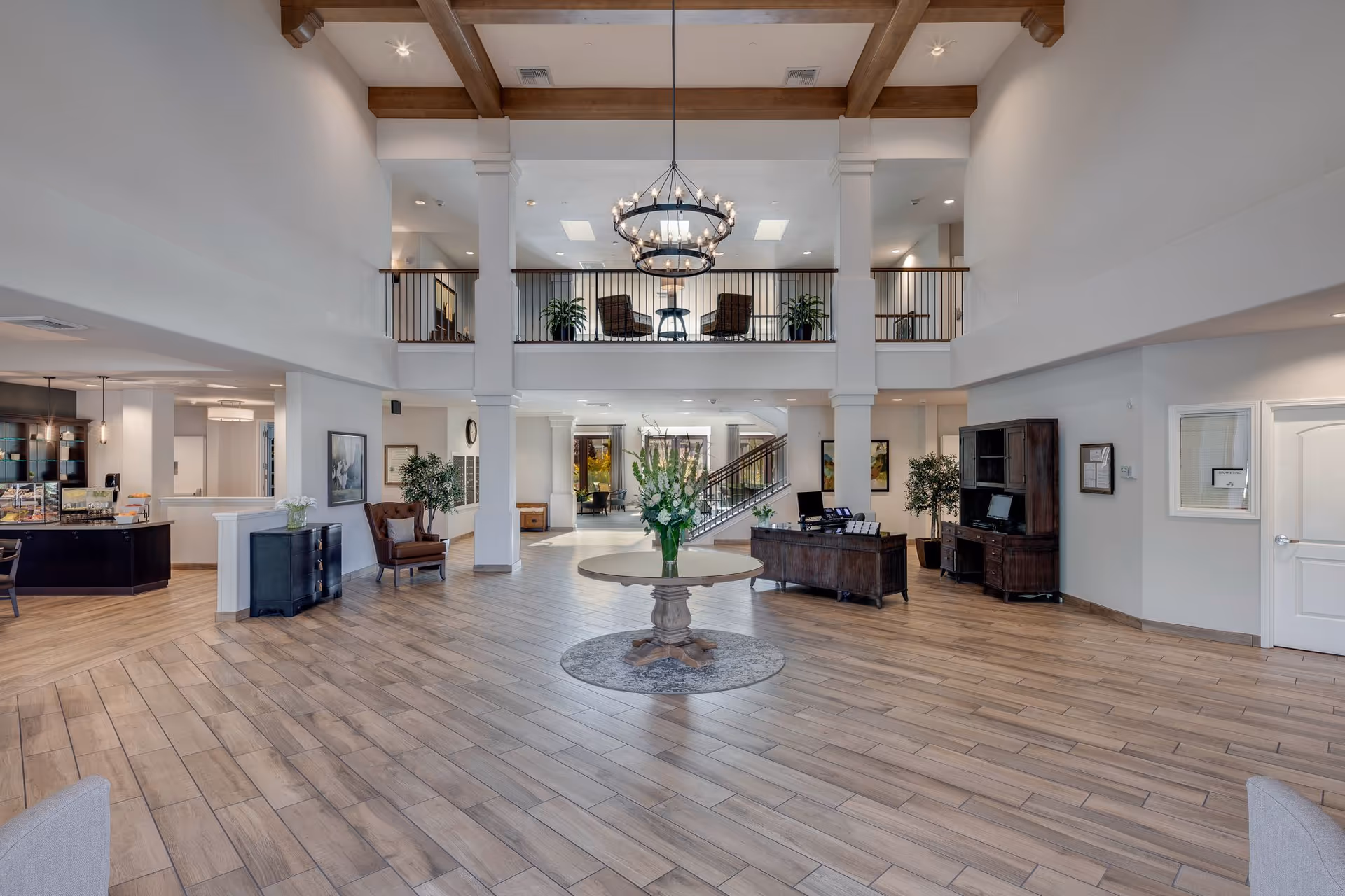 Spacious and well-lit senior living facility lobby with high ceilings, wooden beams, a large chandelier, and a round table with a flower arrangement in the center. The area features wood-style flooring, comfortable seating, plants, and a reception desk. There is a second-floor balcony with chairs and plants overlooking the lobby.
