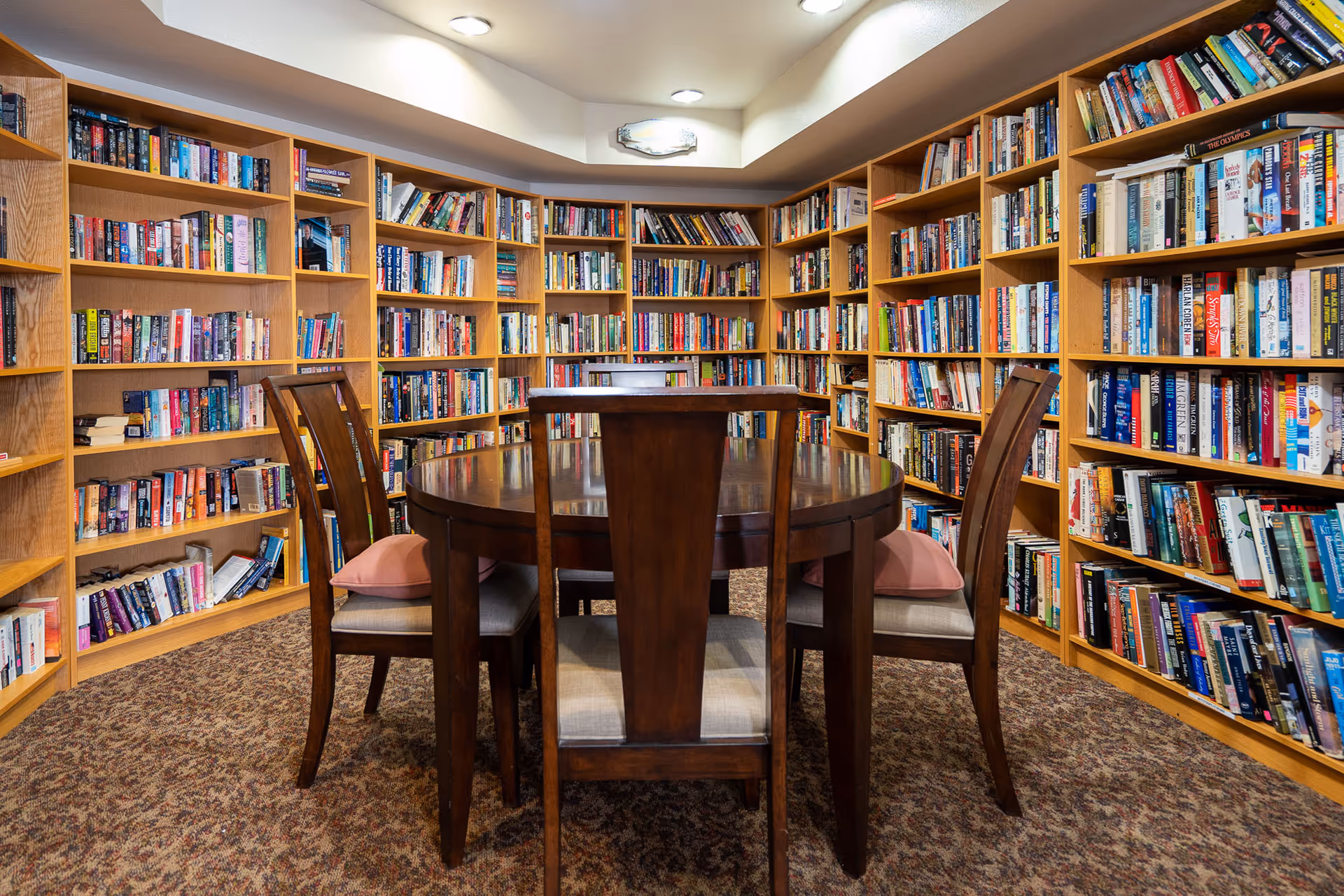 A cozy library room with wooden bookshelves filled with books lining the walls. In the center, there is a round wooden table surrounded by four wooden chairs with cushioned seats. The room has carpeted flooring and a ceiling light fixture.