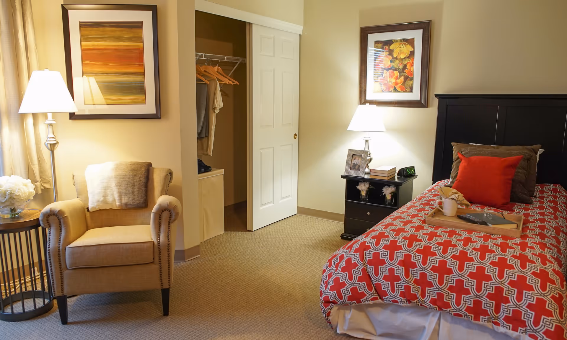 A cozy bedroom in a senior living facility with a single bed covered in a red patterned bedspread, a bedside table with a lamp, books, a clock, and a framed photo. There is a beige armchair with a throw blanket next to a small round table holding a vase of white flowers. The room has warm lighting, framed artwork on the walls, and an open closet with clothes hanging inside.