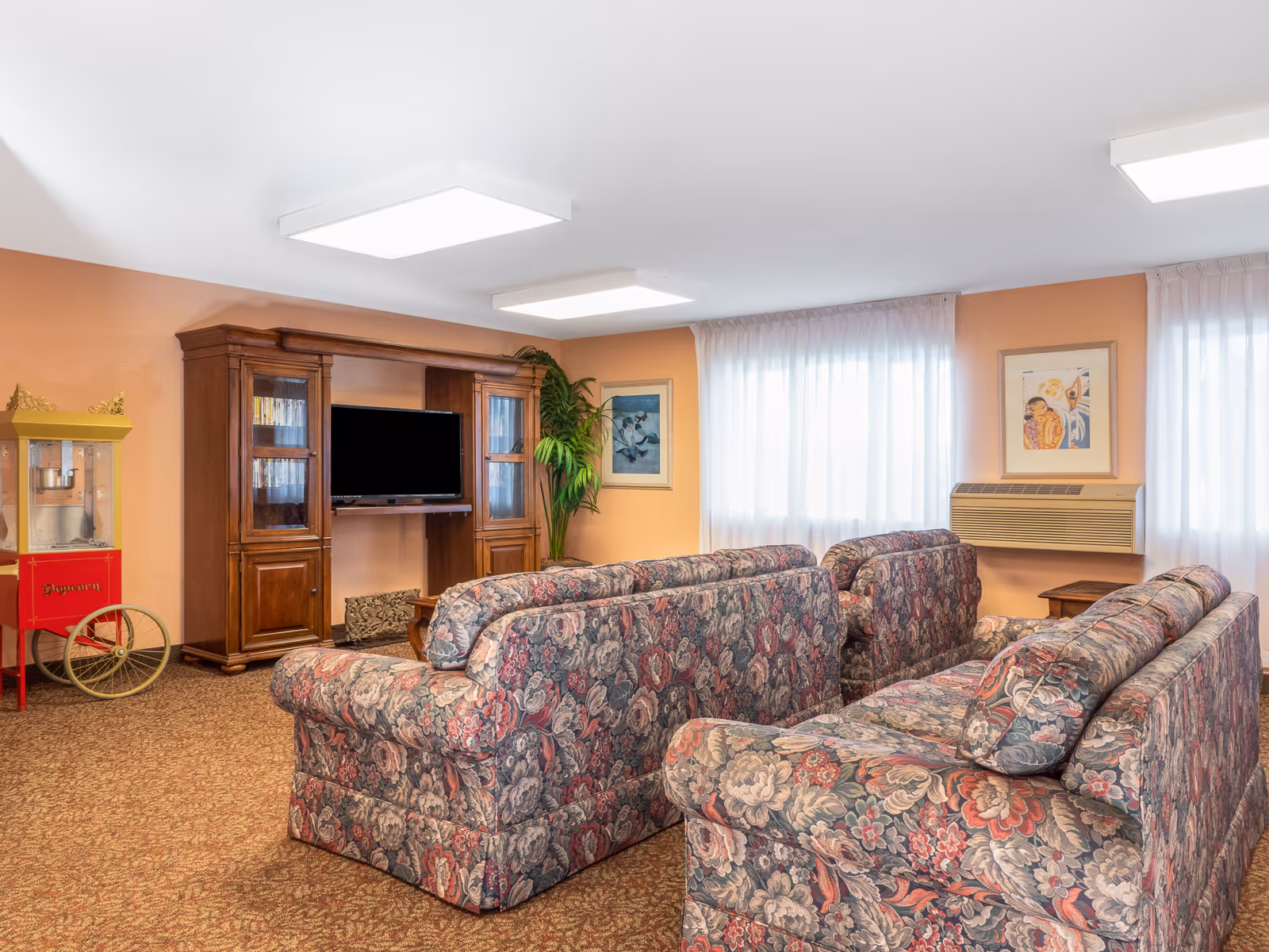 Community living room with patterned floral sofas arranged toward a wooden entertainment center with a TV, a popcorn cart, and framed artwork.