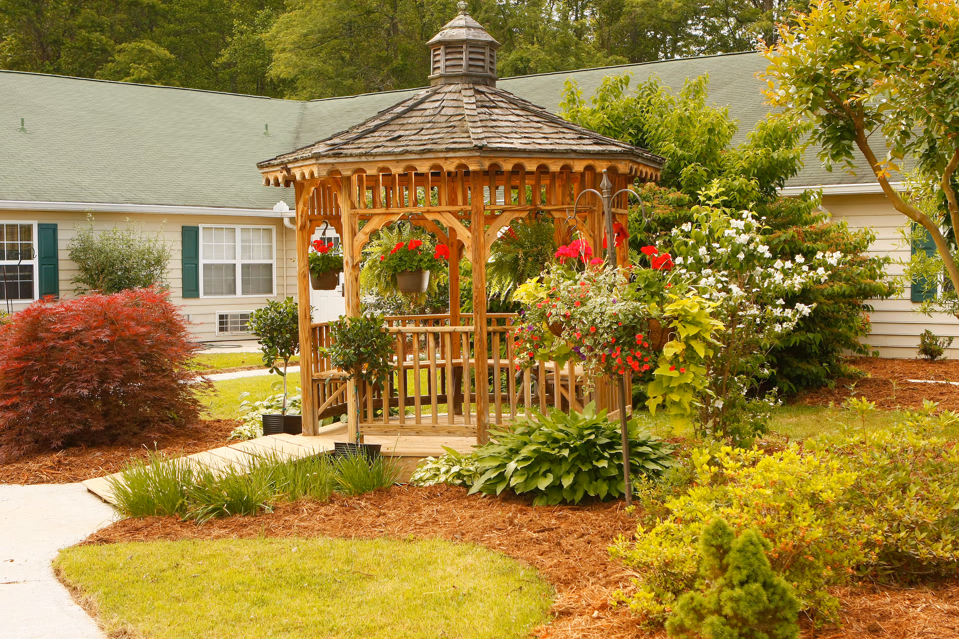 A wooden gazebo surrounded by lush greenery and colorful flowers in the garden area of a senior living facility. The building with green shutters and a green roof is visible in the background.