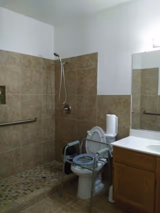 Bathroom with beige tiled walls and floor, featuring a walk-in shower with a handheld showerhead and a grab bar. There is a toilet with a raised seat and arm supports for accessibility, next to a wooden vanity with a white countertop and a large mirror above it.