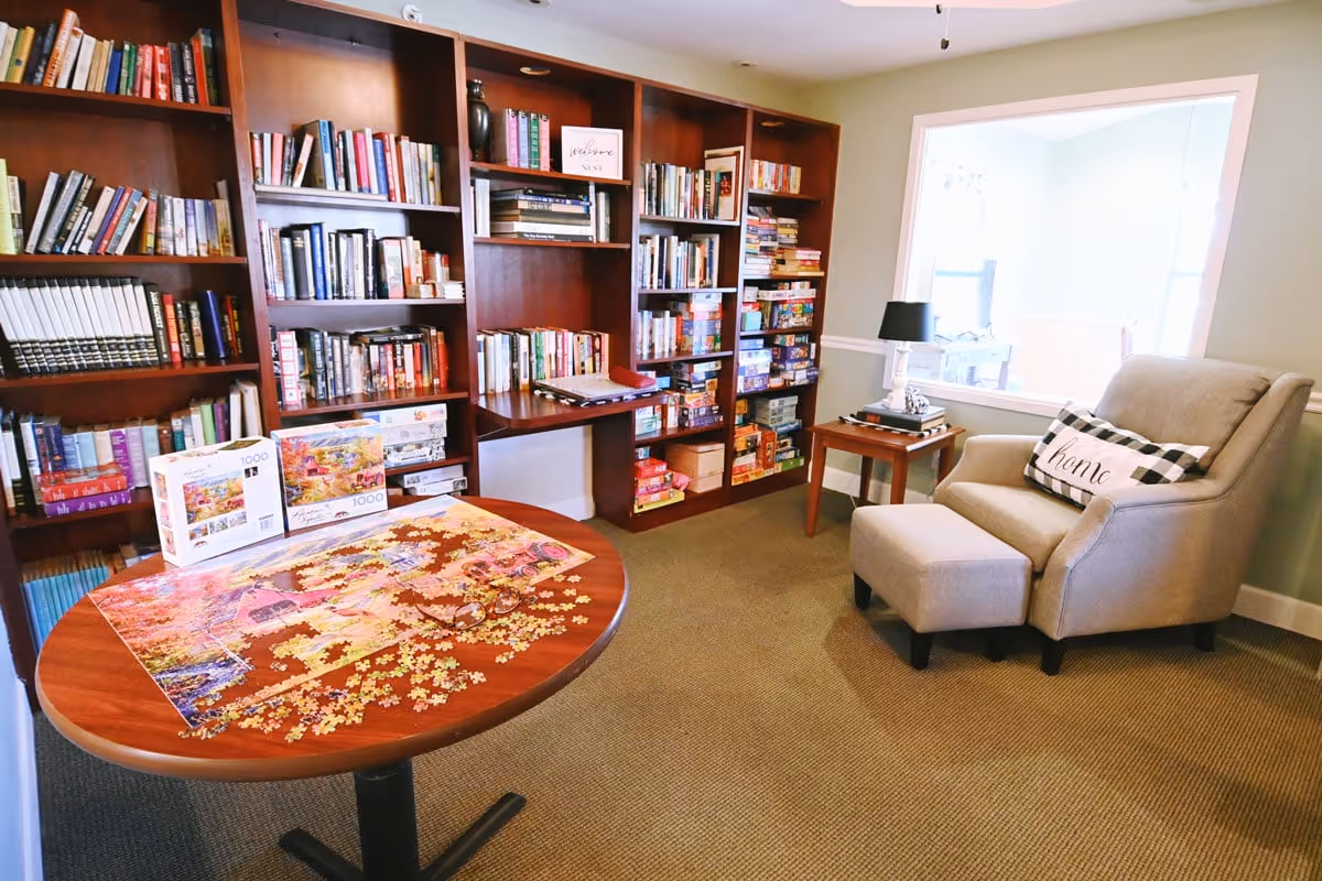 A cozy reading and puzzle room with a large wooden bookshelf filled with books and board games, a round table with an incomplete jigsaw puzzle and puzzle boxes on it, and a comfortable armchair with an ottoman and a decorative pillow that says 'home'. A small side table with a lamp and books is next to the armchair. There is a large window opening to another room.