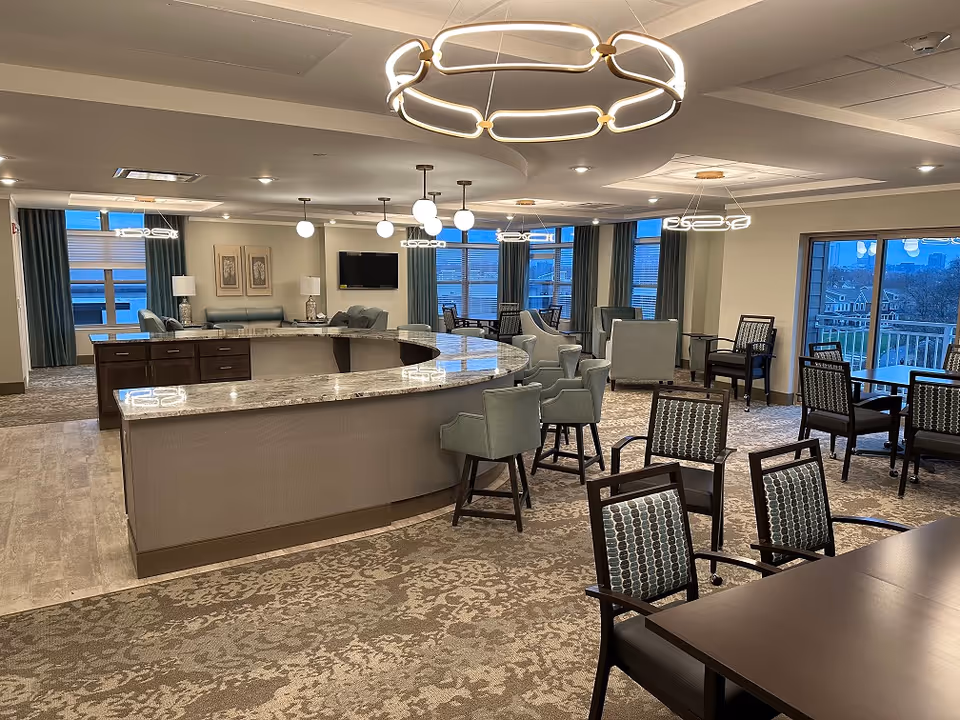 A spacious and well-lit common area in a senior living facility featuring a large curved marble countertop with several upholstered chairs around it. The room has multiple seating areas with armchairs and tables, large windows with blue curtains offering a view outside, modern circular and globe pendant lights hanging from the ceiling, and a wall-mounted flat-screen TV.