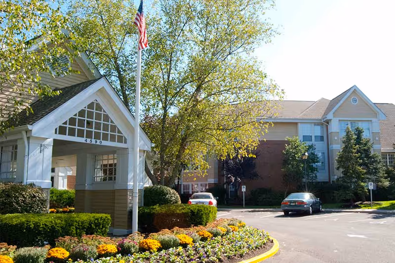 Exterior view of The Forum At Knightsbridge senior living facility showing the entrance with a covered drop-off area, an American flag on a flagpole, landscaped flower beds with colorful flowers, trees, and parked cars in the driveway.
