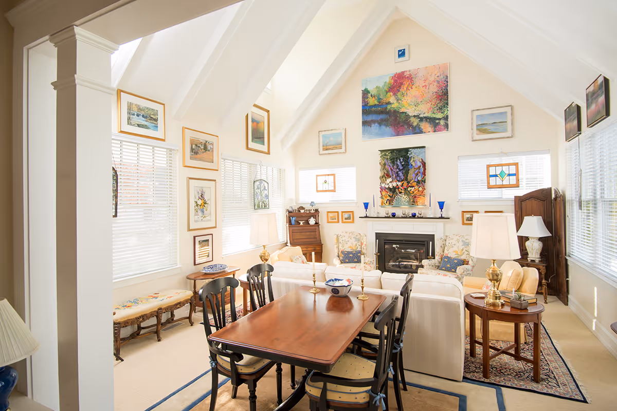 Bright and cozy living room with vaulted ceilings and white beams, featuring a wooden dining table with six chairs in the foreground, two white sofas with floral cushions around a fireplace, various framed paintings and stained glass art on the walls, and multiple lamps providing warm lighting.
