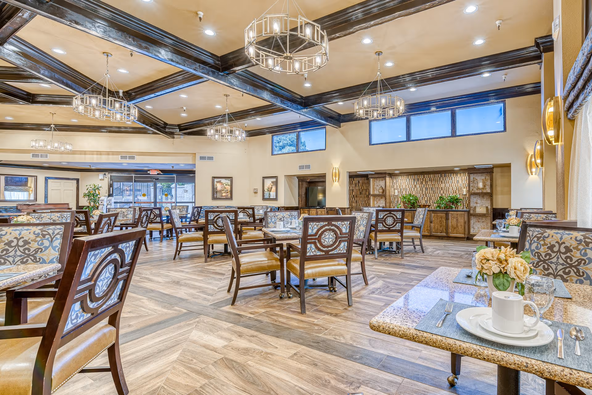 A spacious and well-lit dining room with multiple tables and chairs arranged neatly. The room features wooden flooring, decorative ceiling beams, and modern chandeliers. Tables are set with placemats, plates, cups, and cutlery. There are large windows letting in natural light and some framed artwork on the walls. A sideboard with plants and decorative items is visible in the background.