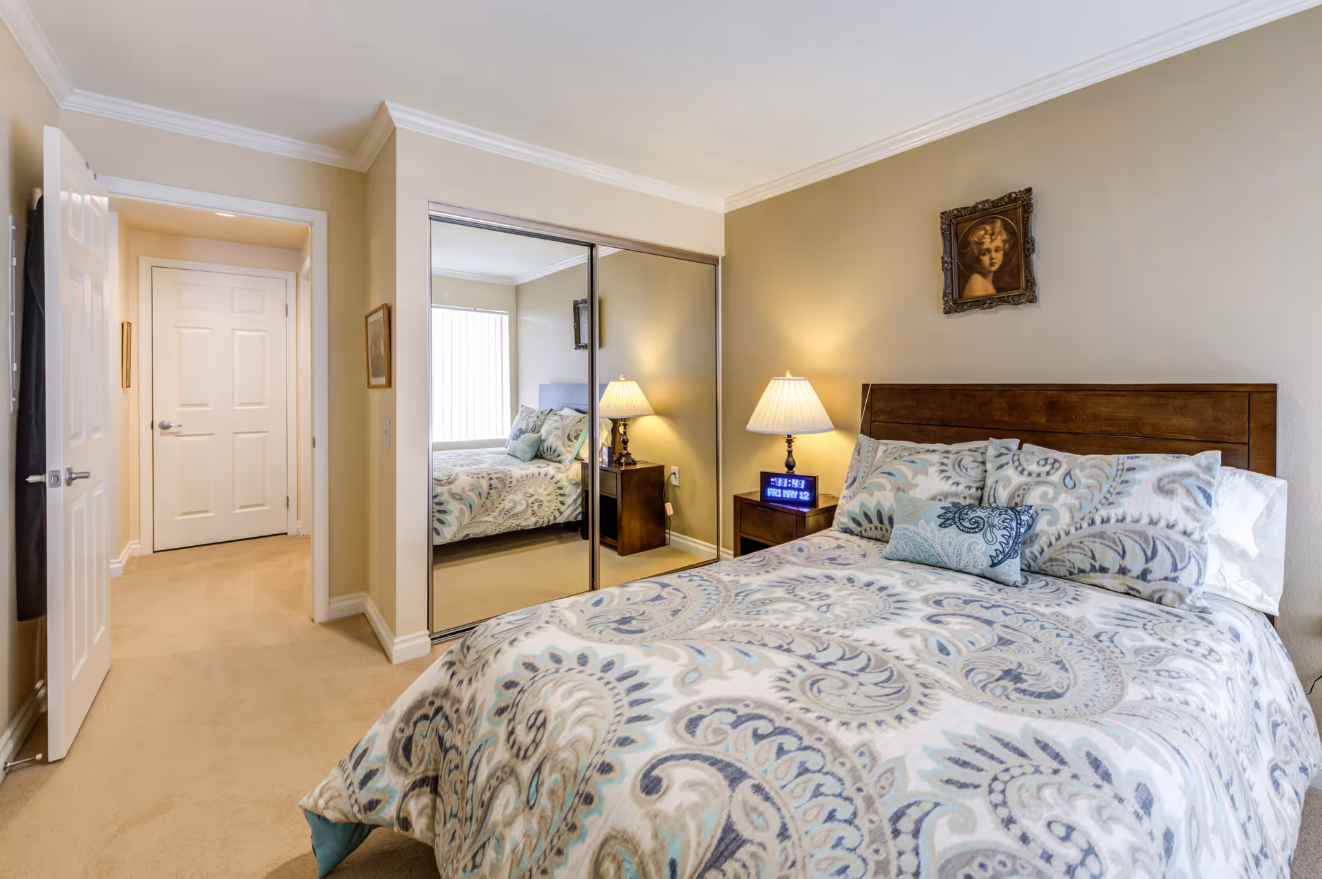 A cozy bedroom with a bed covered in a patterned comforter and multiple pillows. There is a wooden headboard, a nightstand with a lamp and a digital clock, and a framed portrait hanging on the beige wall. A mirrored sliding closet door reflects part of the bed and window with vertical blinds. The room has beige carpet and light-colored walls with white trim.