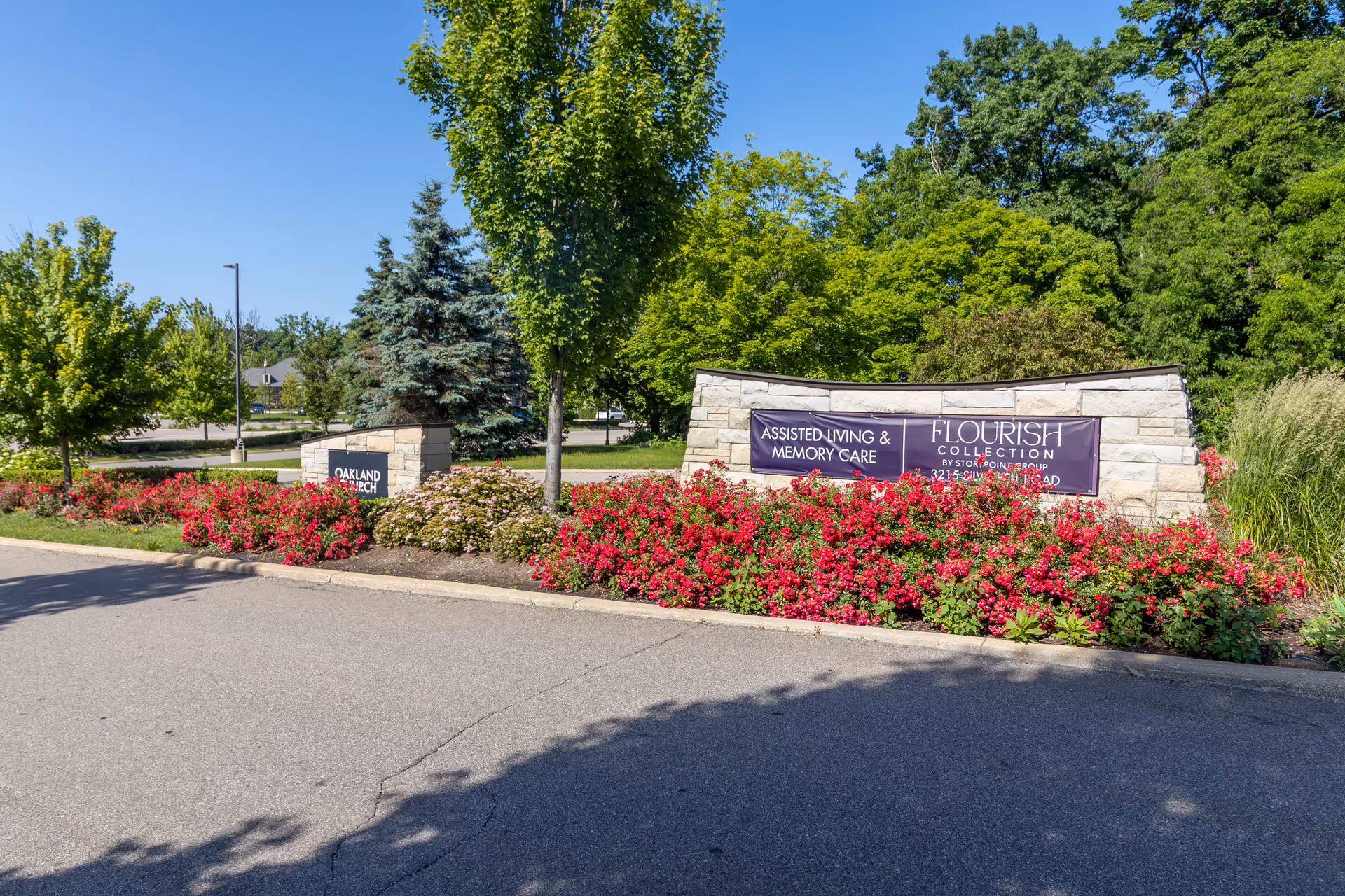 Stone entrance sign for the Flourish Collection surrounded by red flowers, trees, and landscaping under a clear blue sky.