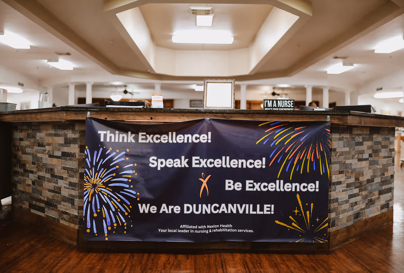 Reception desk area inside Duncanville Healthcare & Rehabilitation Center with a banner hanging on the front that reads 'Think Excellence! Speak Excellence! Be Excellence! We Are DUNCANVILLE!' The desk has a stone tile facade and there is a sign on top that says 'I'M A NURSE'.