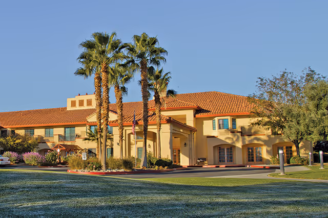 Two-story stucco building with a red tile roof, palm trees and a circular driveway on a manicured lawn under a clear blue sky.