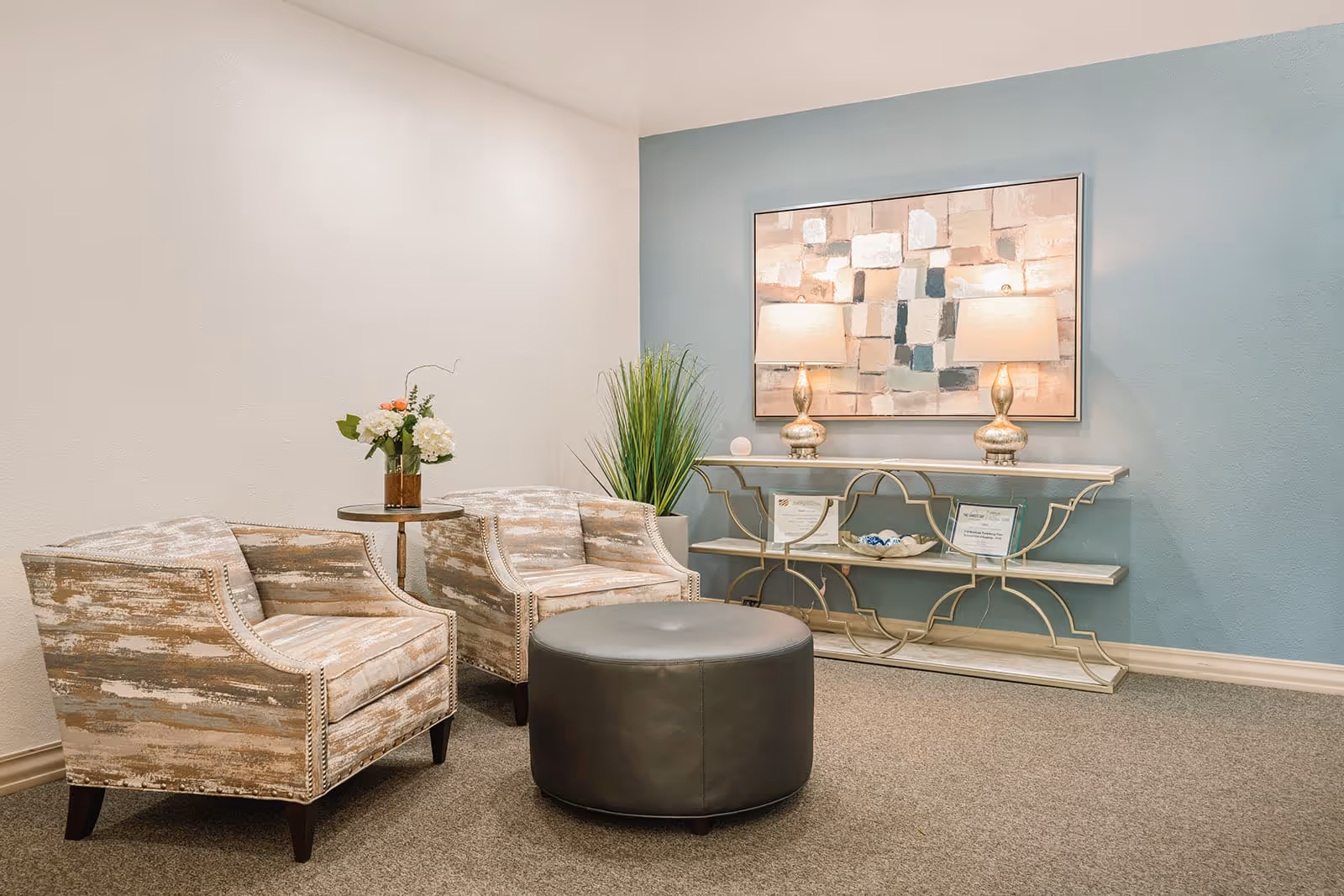 A cozy sitting area with two patterned armchairs, a round gray ottoman, and a small round side table with a vase of flowers. Behind the chairs is a decorative console table with two lamps and framed certificates, set against a blue accent wall with an abstract painting above.