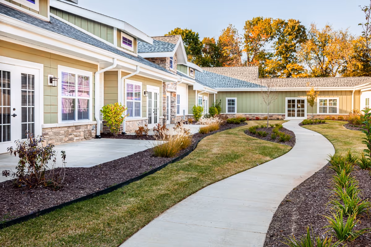 Curved concrete walkway leading through landscaped garden beds with shrubs and small trees, alongside a single-story building with green siding, stone accents, and multiple windows and doors, under a clear sky with autumn-colored trees in the background.