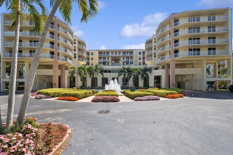 Front exterior view of a multi-story senior living facility named Park Summit, featuring balconies, palm trees, a central water fountain, and landscaped flower beds under a blue sky with some clouds.