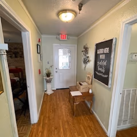 A hallway with wooden flooring leading to a white door with a glass window. On the right wall, there is a decorative wall hanging and a blackboard with white text. A small wooden table with a box and some papers is placed against the right wall. The left side shows an open doorway leading to another room with a bed and some furniture visible. The ceiling has a light fixture and an exit sign above the door.