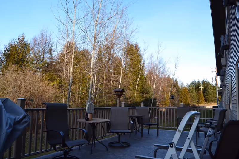 Outdoor patio area with several chairs and small tables, surrounded by a wooden railing. The patio overlooks a wooded area with trees and bushes under a clear blue sky.