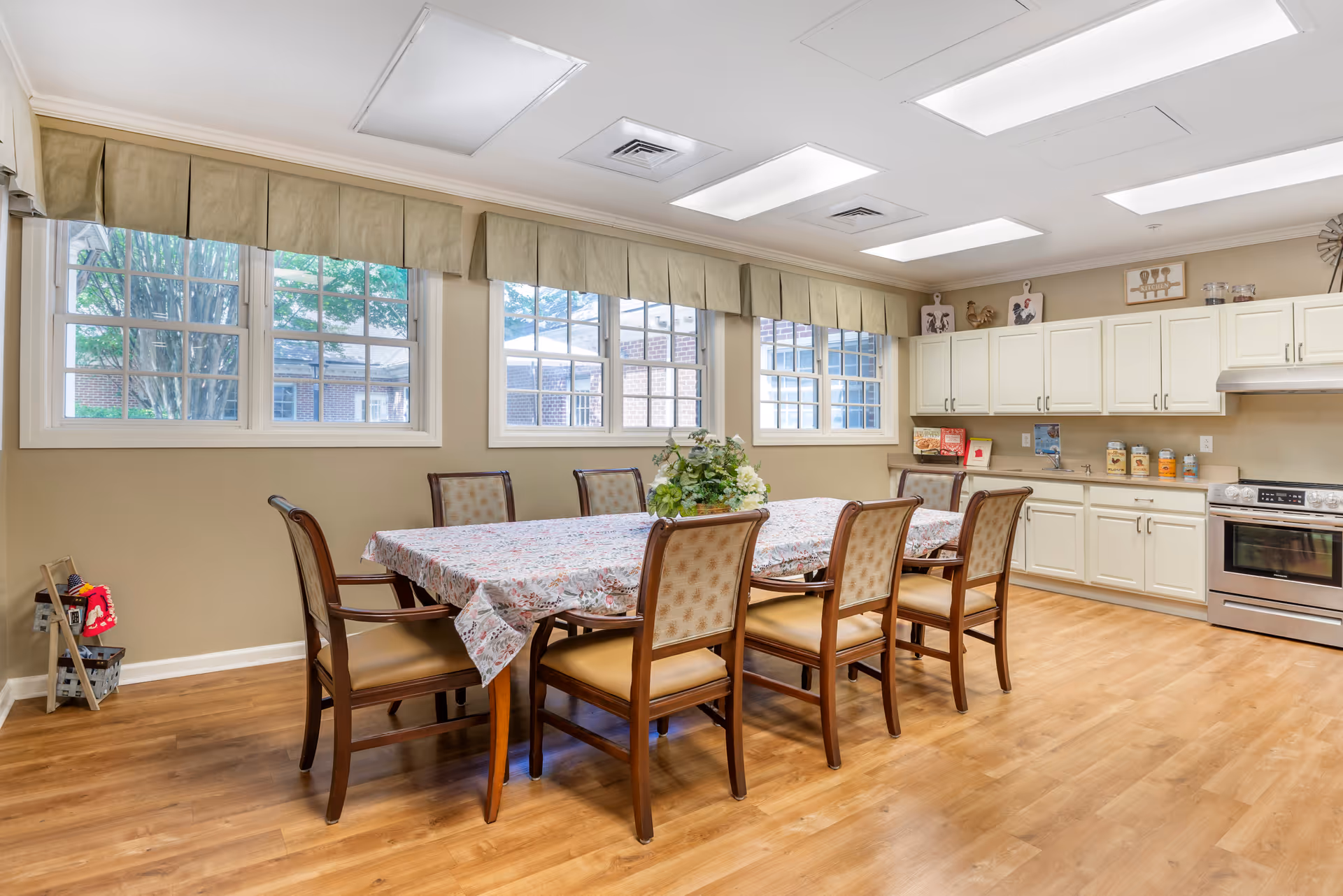 A bright and spacious kitchen and dining area with a long table covered by a floral tablecloth surrounded by eight wooden chairs with cushioned seats. The room has large windows with beige valances, white cabinetry, a stainless steel oven, and various kitchen decorations on the countertops and walls. The floor is wooden, and the ceiling has multiple fluorescent light panels.