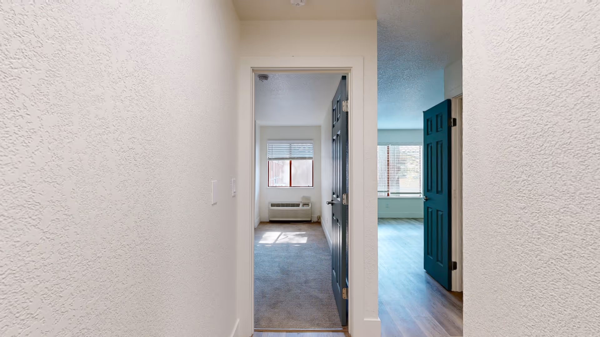 View down a hallway with textured white walls leading to two open doorways. The left doorway reveals a small carpeted room with a window and an air conditioning unit beneath it. The right doorway opens into a larger room with wood flooring and two windows with blinds, letting in natural light.