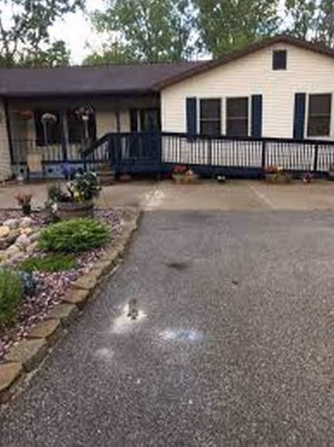 Exterior view of a single-story building with white siding and dark blue trim, featuring a ramp and stairs leading to the entrance. There is a paved driveway in front and a landscaped garden area with rocks and plants on the left side.