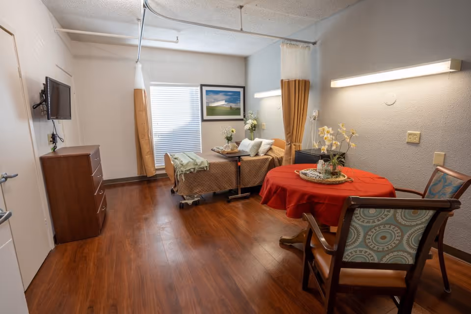 Assisted-living bedroom with a single hospital bed, round table and chairs, dresser and wall-mounted TV in a warmly lit room.