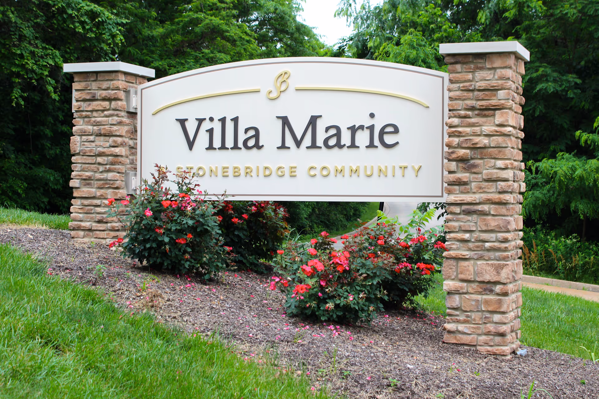 Entrance sign reading 'Villa Marie' mounted between two stone pillars with surrounding flowers and trees.