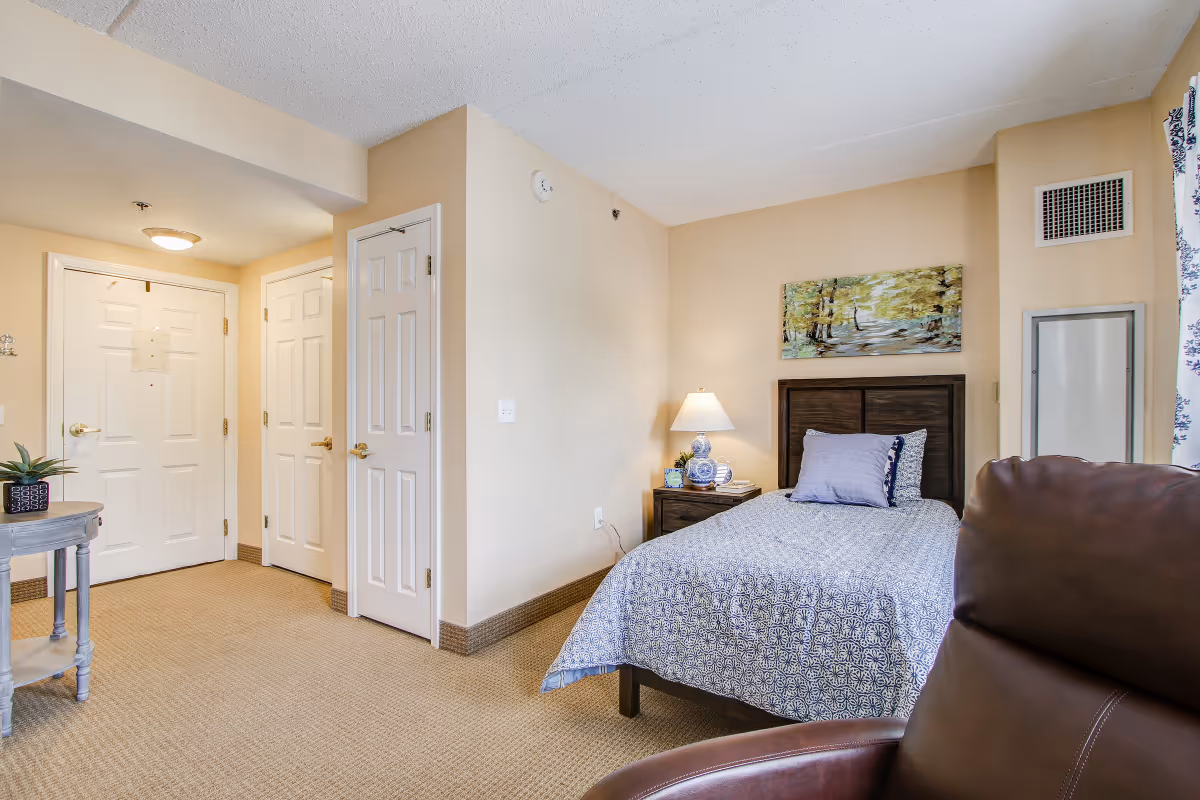 A cozy bedroom in a senior living facility featuring a single bed with patterned bedding and pillows, a wooden nightstand with a lamp and decorative items, a painting of a forest scene on the wall above the bed, a brown leather chair in the foreground, and a small round table with a plant near the entrance door. The room has beige walls and carpeted flooring.