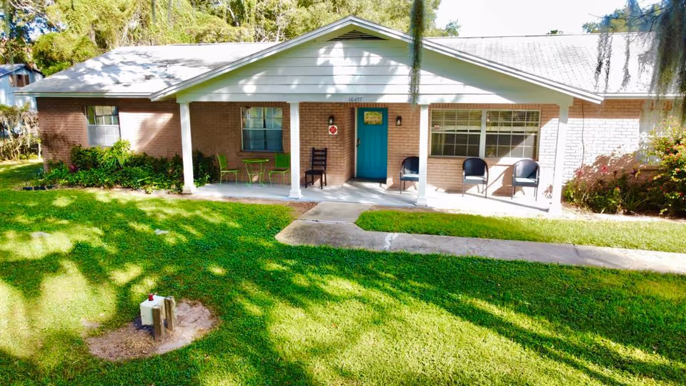 Front exterior view of a single-story brick building with a white roof and a covered porch. The porch has a turquoise door, two black chairs, a wooden chair, and a small green table with two green chairs. The building is surrounded by green grass and some bushes, with trees casting shadows on the lawn.