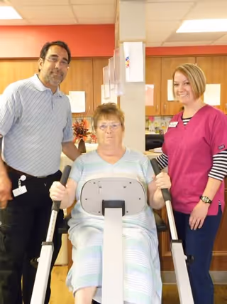 A woman seated on a piece of exercise equipment in a room with wooden cabinets, flanked by a man in a light blue polo shirt and a woman in a pink scrub top, both standing and smiling.