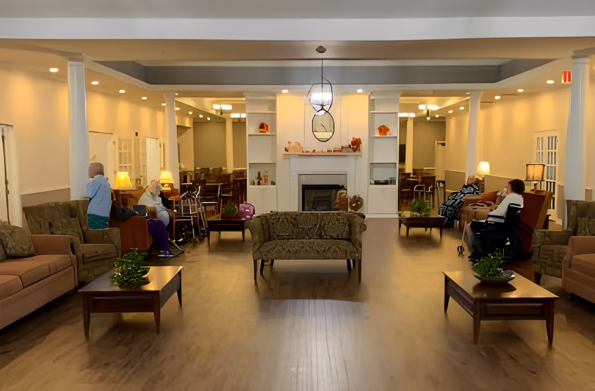 A spacious senior living facility common area with multiple armchairs and sofas arranged around wooden coffee tables with green plants. Several elderly individuals are seated, some in wheelchairs, and there is a fireplace with decorative items on the mantel in the background. The room is warmly lit with lamps and recessed ceiling lights.