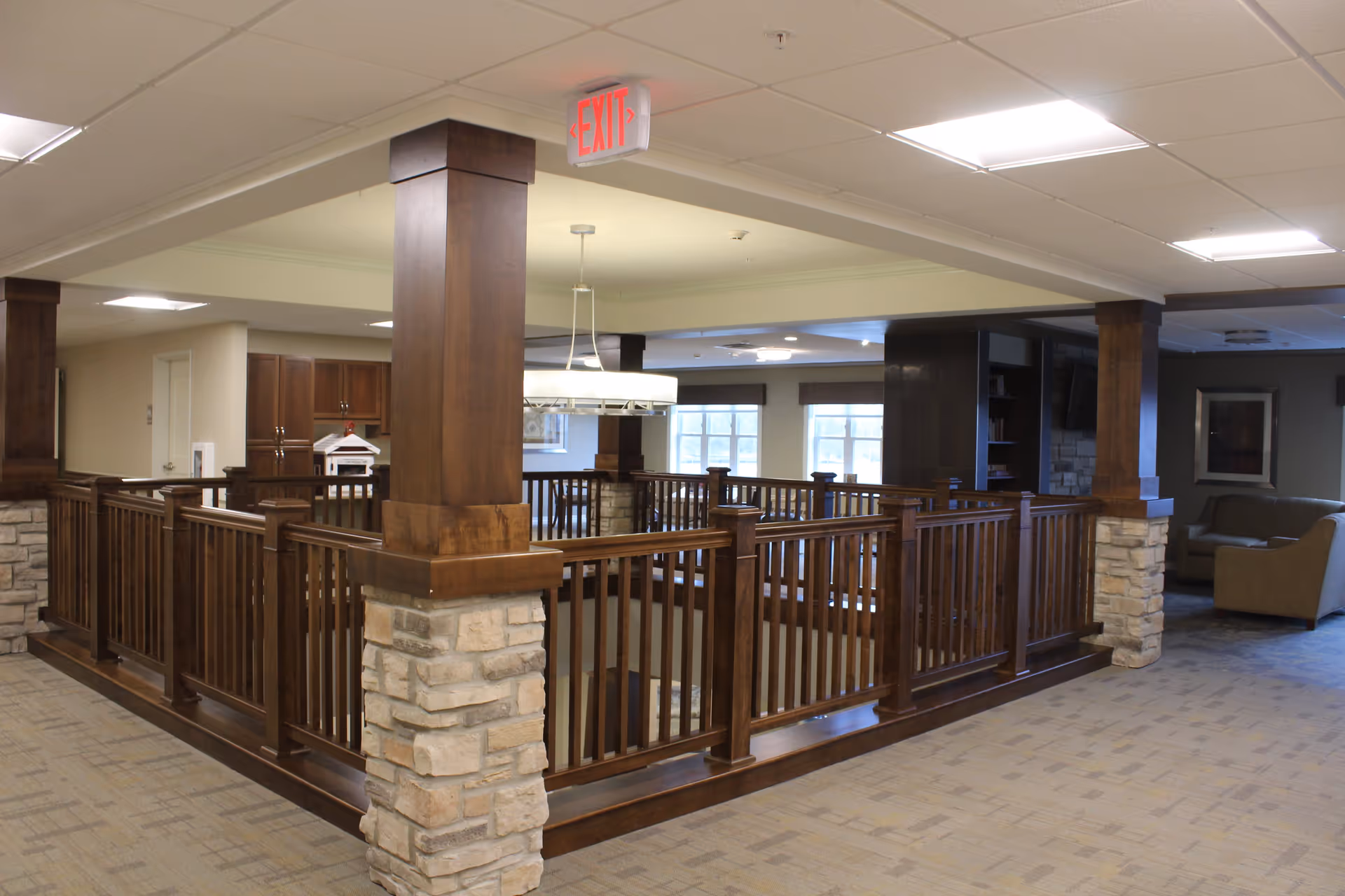 Interior view of a senior living facility showing a spacious common area with wooden railings around a stairwell, stone pillars, carpeted floor, ceiling lights, and seating areas with sofas in the background.