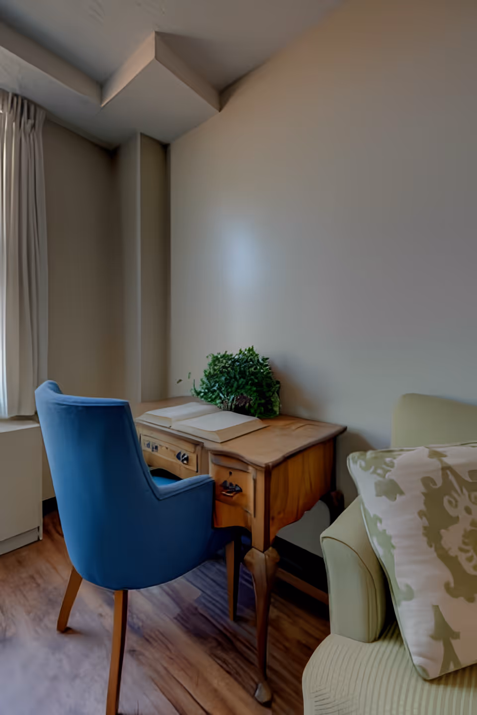 A cozy corner of a room featuring a wooden desk with an open book and a green plant on top. A blue upholstered chair is positioned in front of the desk. To the right, part of a light green armchair with a patterned cushion is visible. The room has wooden flooring and light-colored walls, with a window covered by white curtains on the left side.