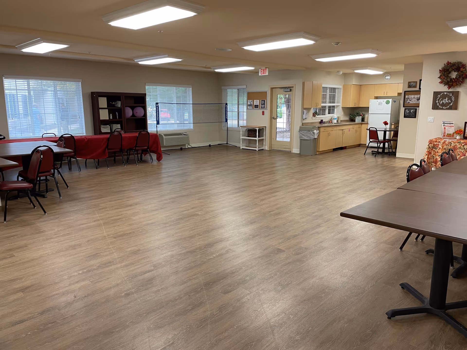 A spacious multipurpose room with wood flooring and fluorescent ceiling lights. The room contains several tables and chairs arranged along the walls, a small kitchen area with light wood cabinets, a refrigerator, and a sink. There are windows with blinds, a door leading outside, and a badminton net set up in the middle of the room. The walls have some decorations including a wreath and framed pictures.