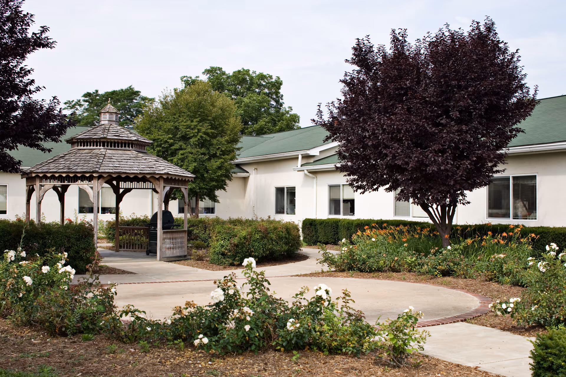 A landscaped courtyard with a wooden gazebo, walkways, flower beds, and a white single-story building in the background.
